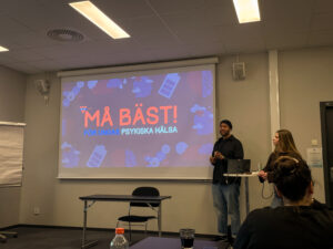 Stephen och Tura står framför en presentation som det står "Må Bäst - för ungas psykiska hälsa" på. 