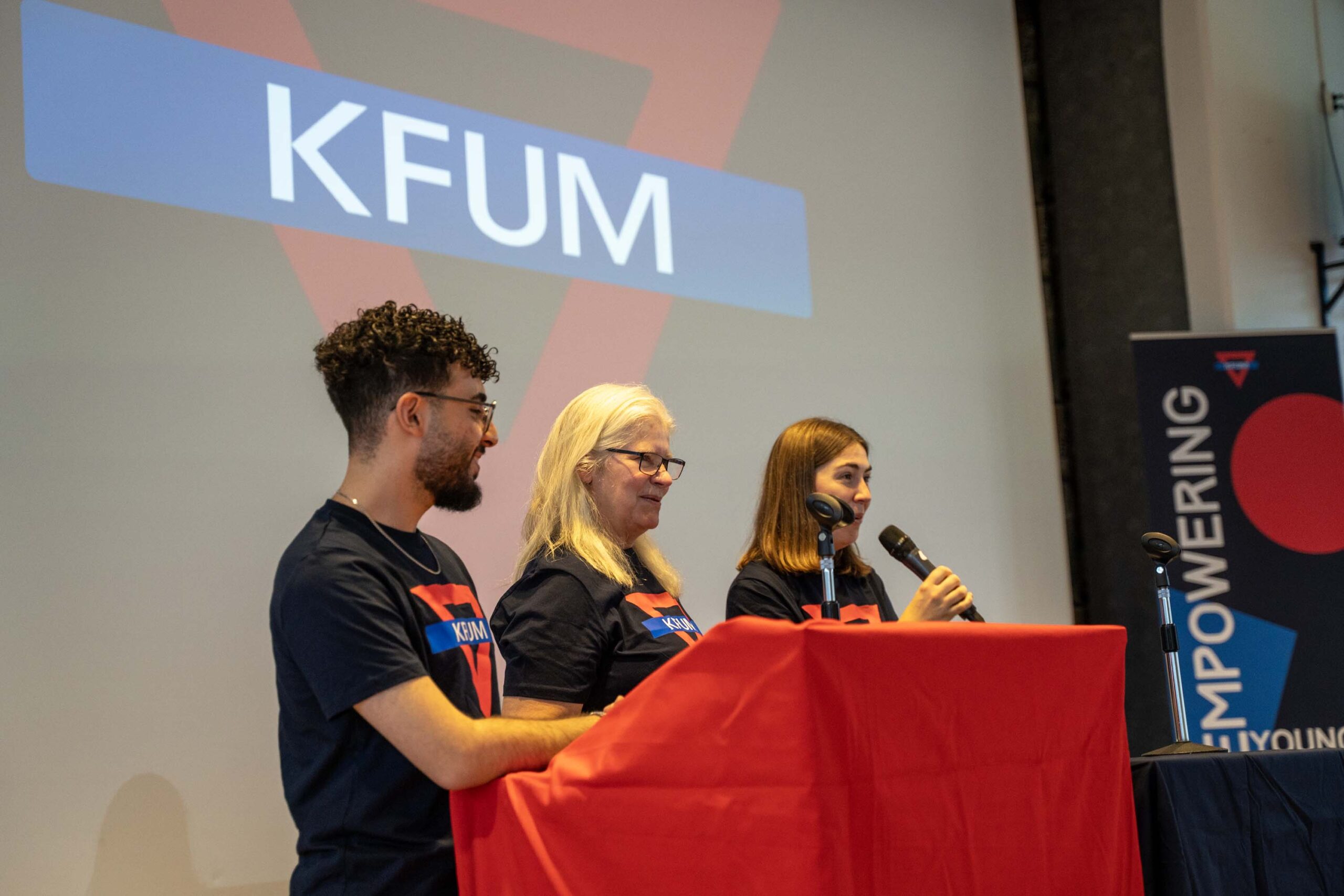 Tre personer som står på scen. En av personerna pratar i en mikrofon. I bakgrunden syns KFUMs logotyp.